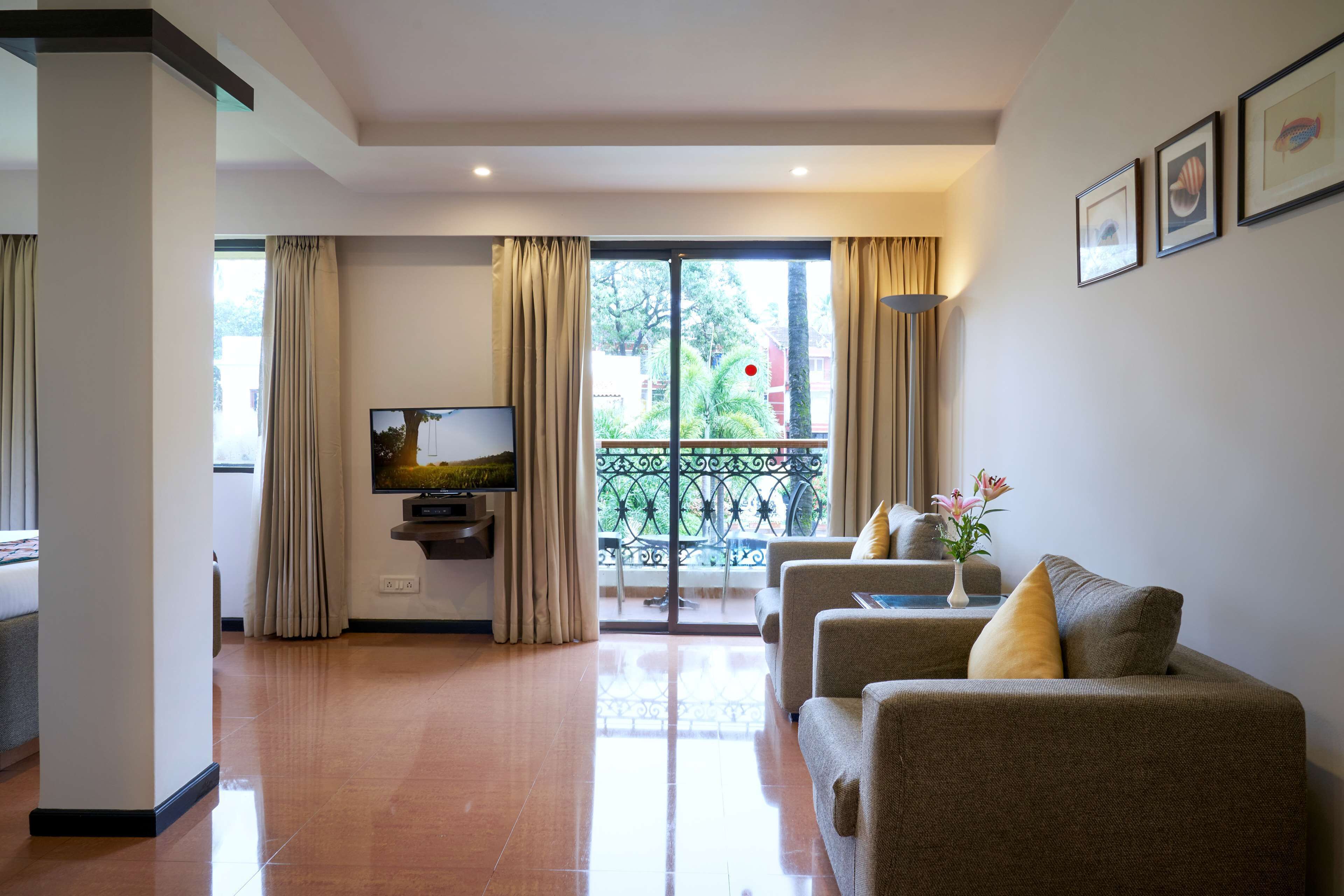 A living room with a sofa set, a flat-screen TV, and a balcony with a view of greenery.