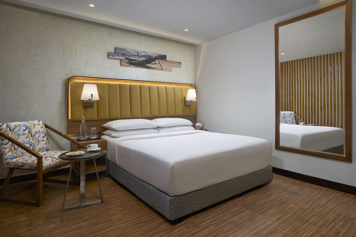 A hotel room with a large bed, a chair, a small table with a cup and saucer, and a wall-mounted lamp.