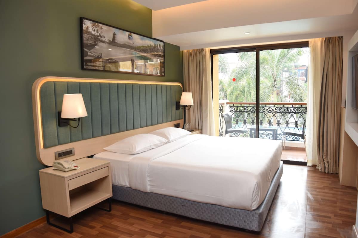 A hotel room with a large bed, two bedside lamps, a nightstand with a phone, a painting on the wall, and a balcony with a view of palm trees.