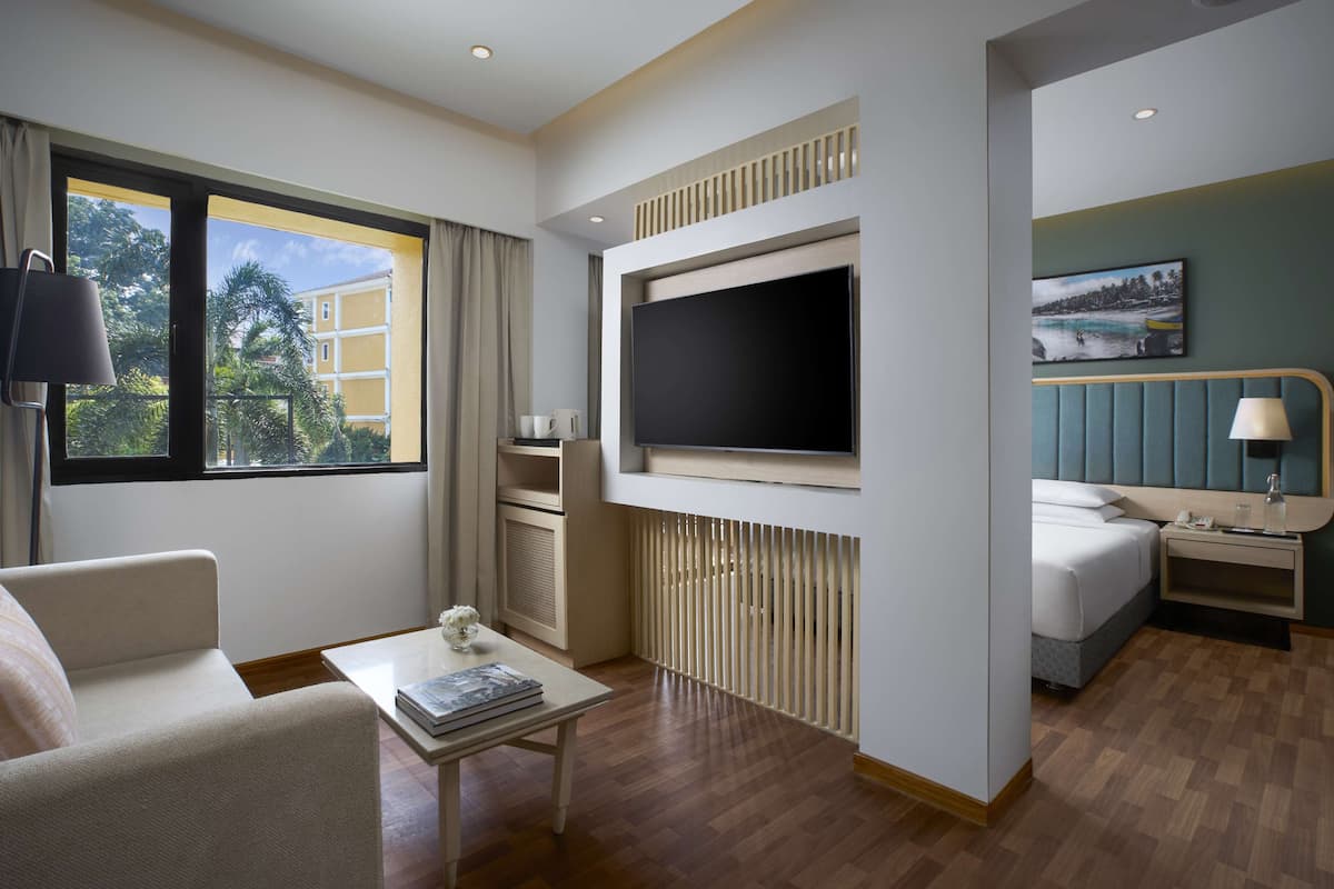 A hotel room with a large flat-screen TV, a bed, a desk with a lamp, a coffee table with a magazine, and a window with a view of greenery and buildings.