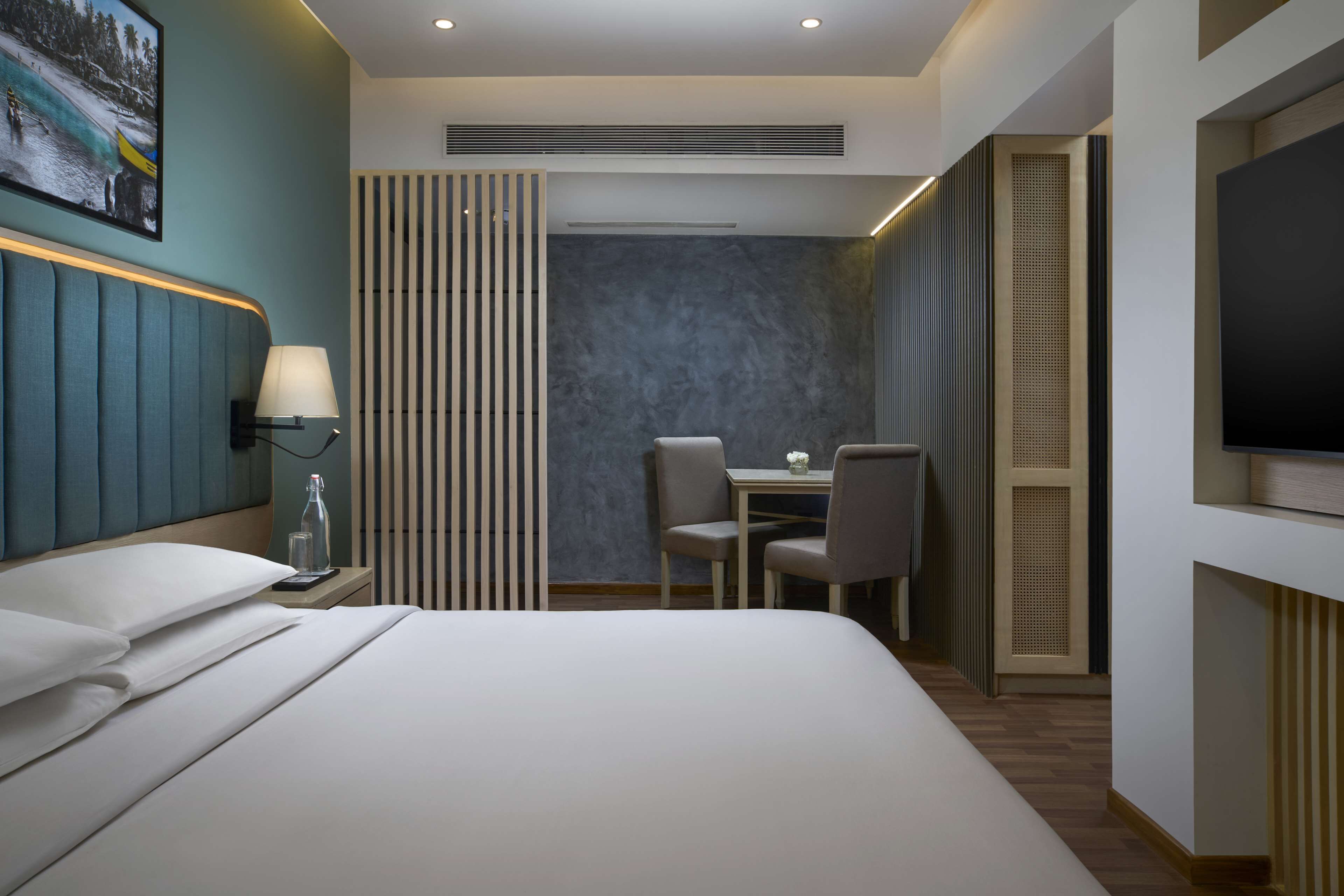 A modern hotel room with a bed, bedside table, lamp, TV, and a small dining area with chairs and a table.