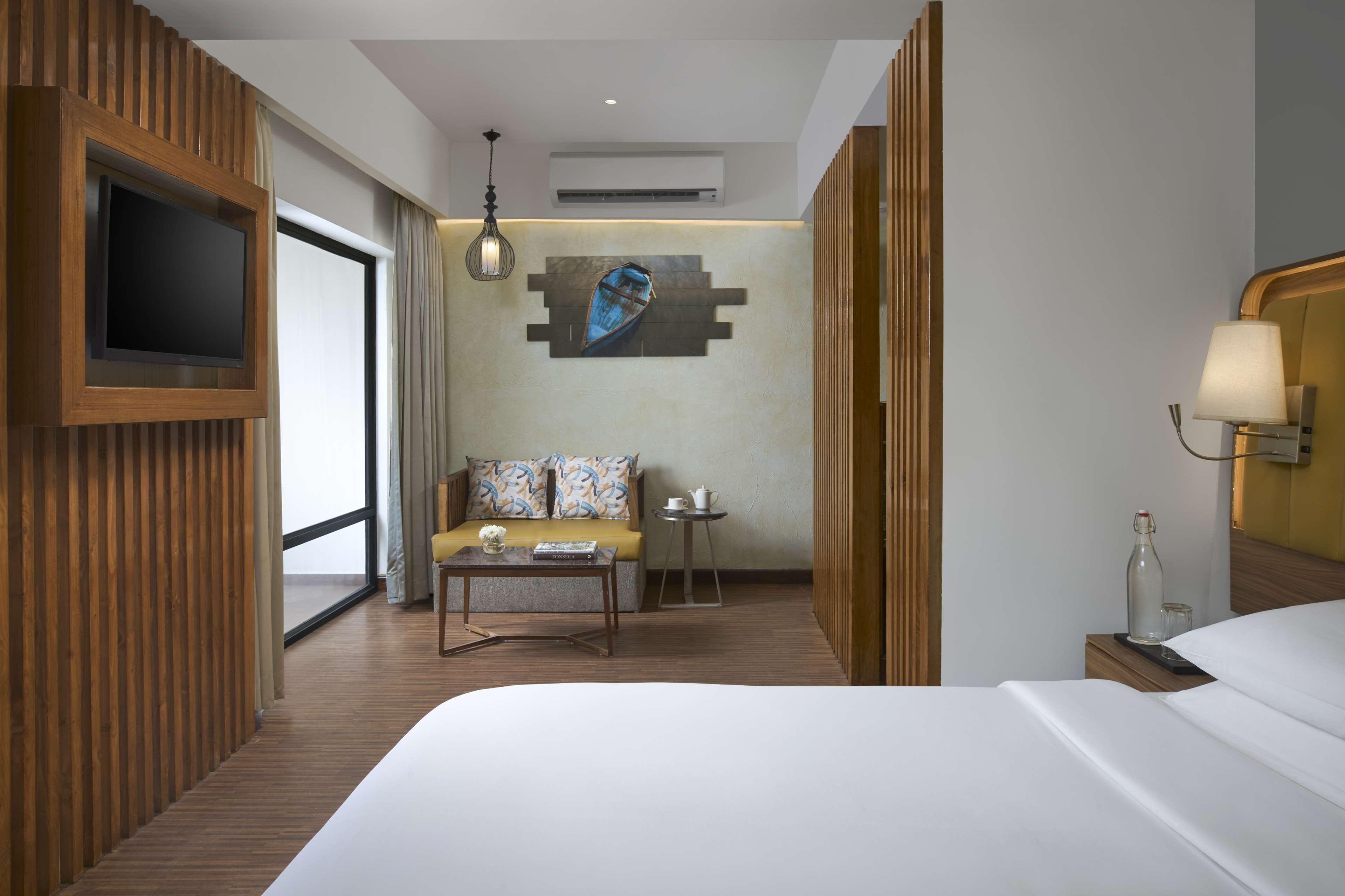 A hotel room with a large bed, a sofa, a coffee table, a TV mounted on the wall, a painting, and a window with curtains.