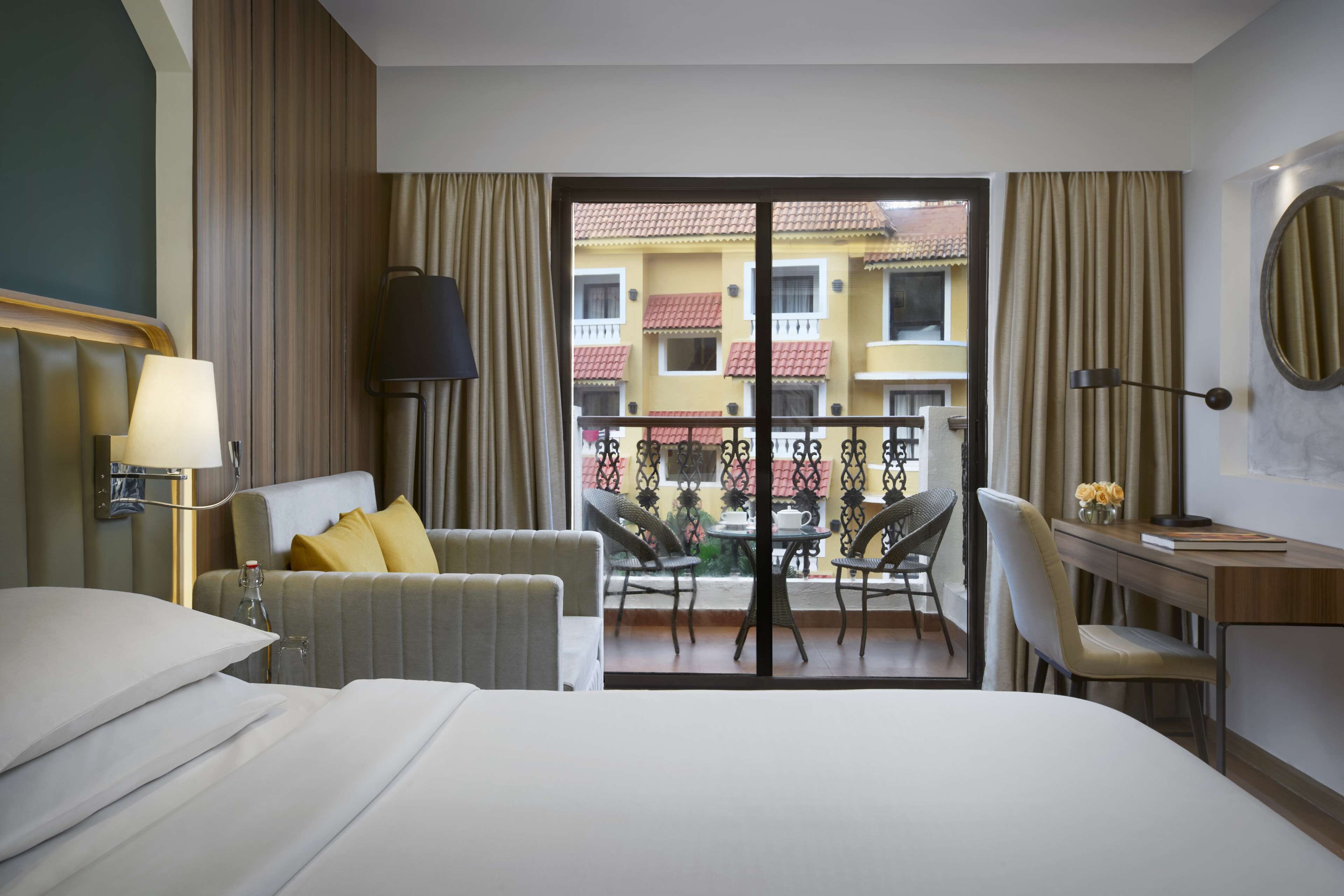 Ein Hotelzimmer mit Bett, Schreibtisch, Stuhl, Tisch mit Stühlen, einem Balkon mit Blick auf Gebäude und einer Lampe.
