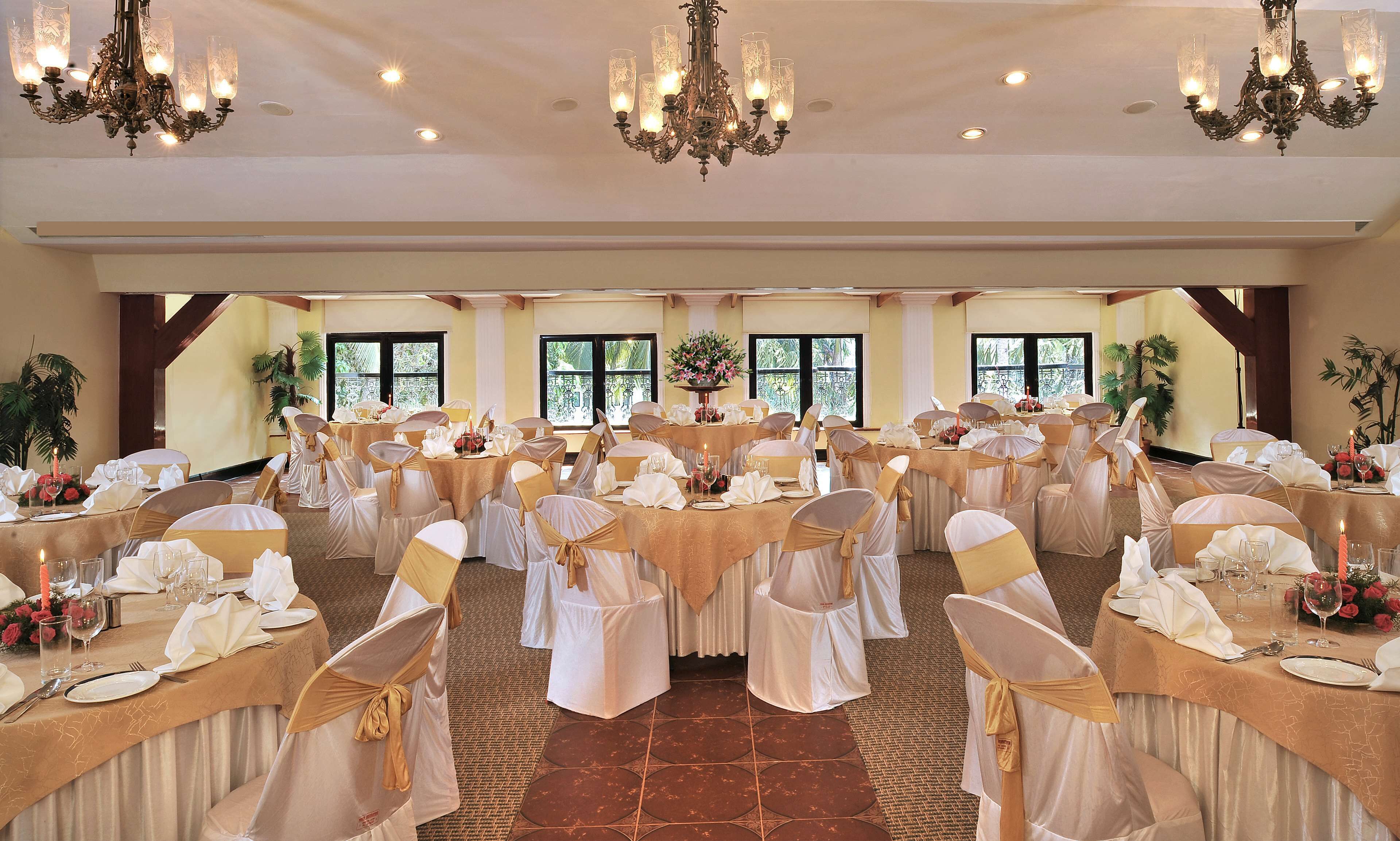 Un salón de banquetes con mesas redondas, sillas con cintas doradas, candelabros y ventanas amplias con vistas.