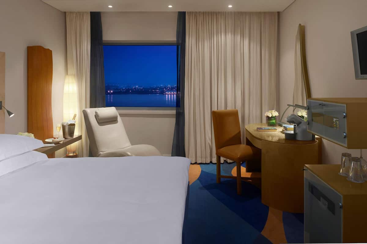 A hotel room with a bed, a desk, a chair, a nightstand, a lamp, and a view of the city at night.