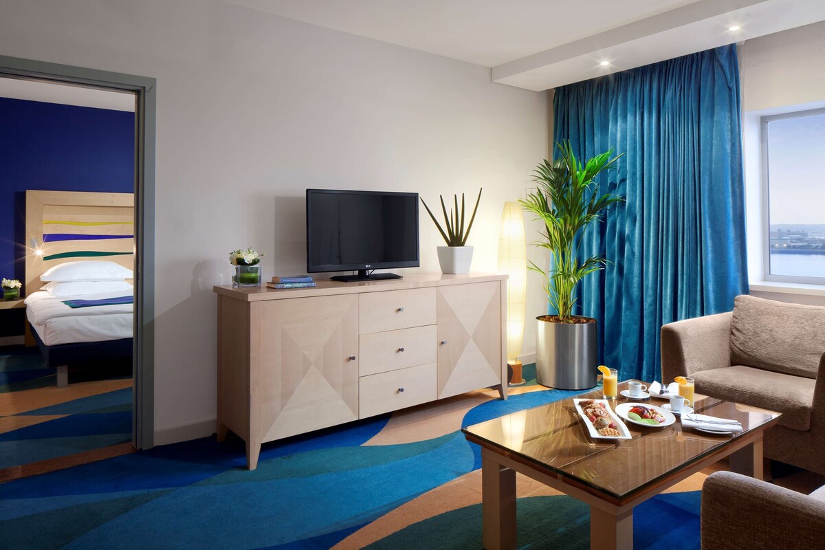 A hotel room with a bed, a TV on a cabinet, a dining table with breakfast, a sofa, and a large window.