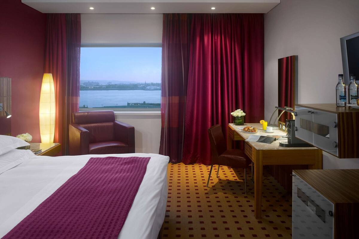 A hotel room with a bed, a desk, a chair, a lamp, a mirror, and a view of a body of water.