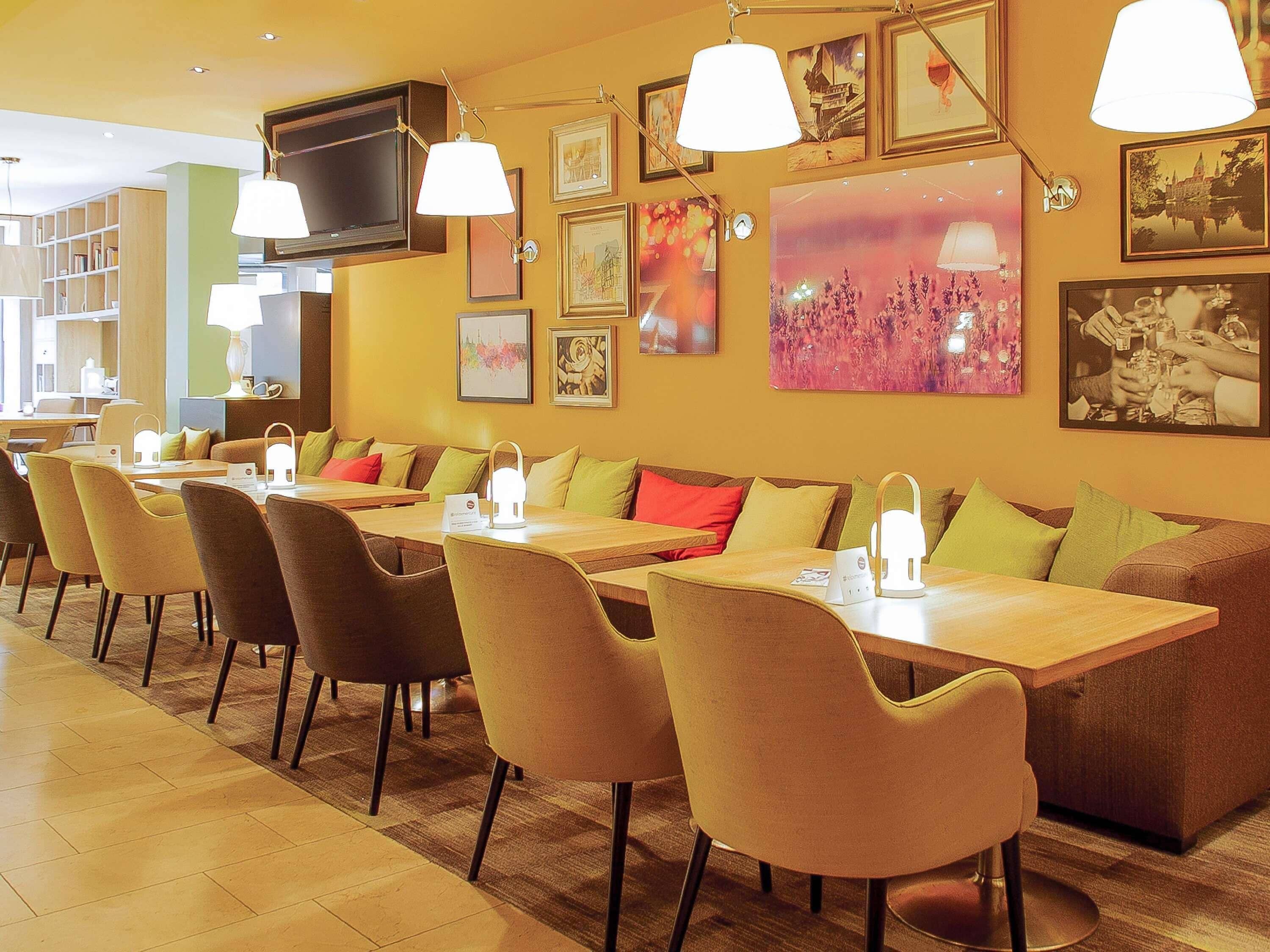 A restaurant with a long dining table, multiple chairs, and a sofa. There are framed pictures on the walls and a television mounted on the wall.