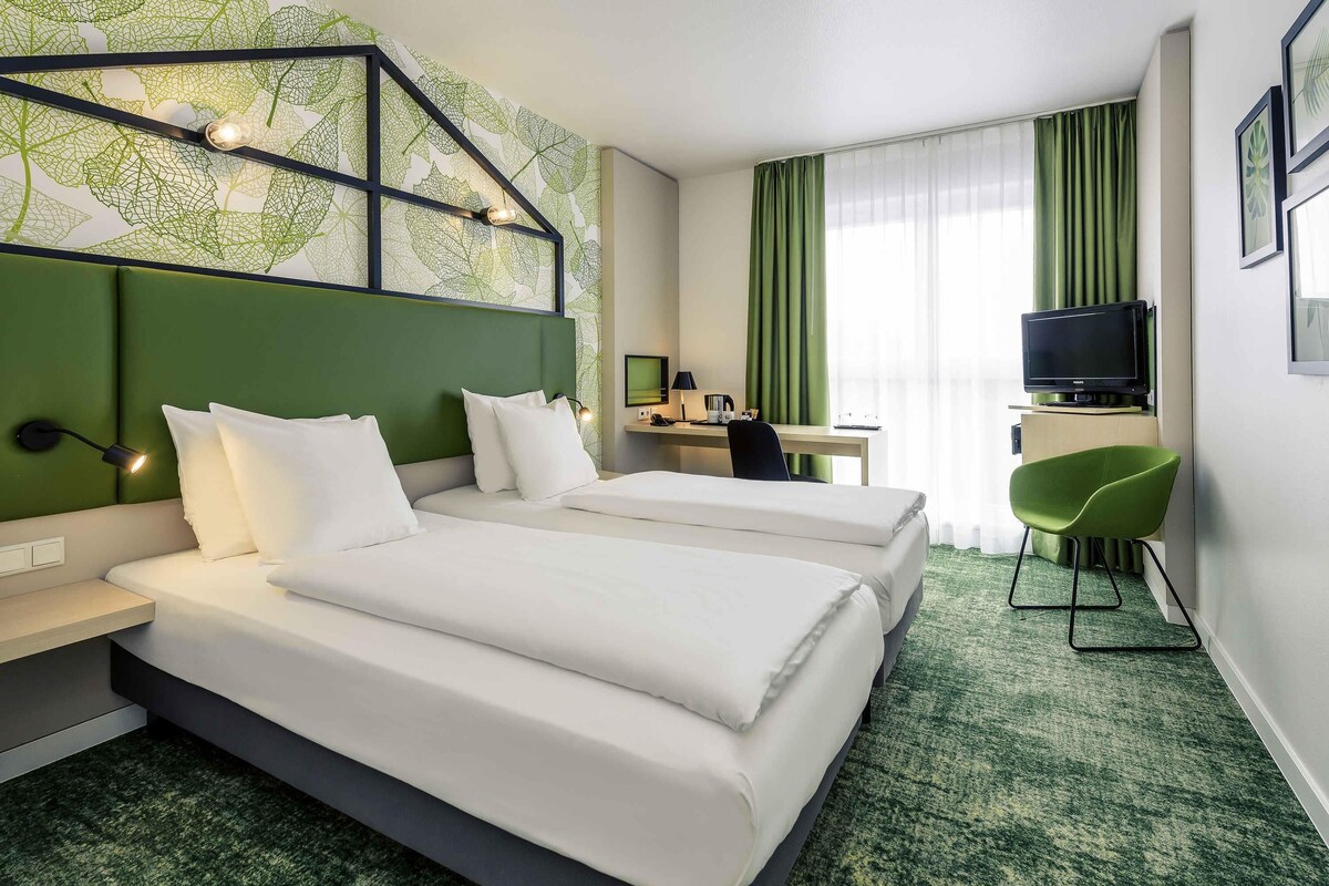 A hotel room with two beds, a desk, a chair, a TV, and a green carpet.