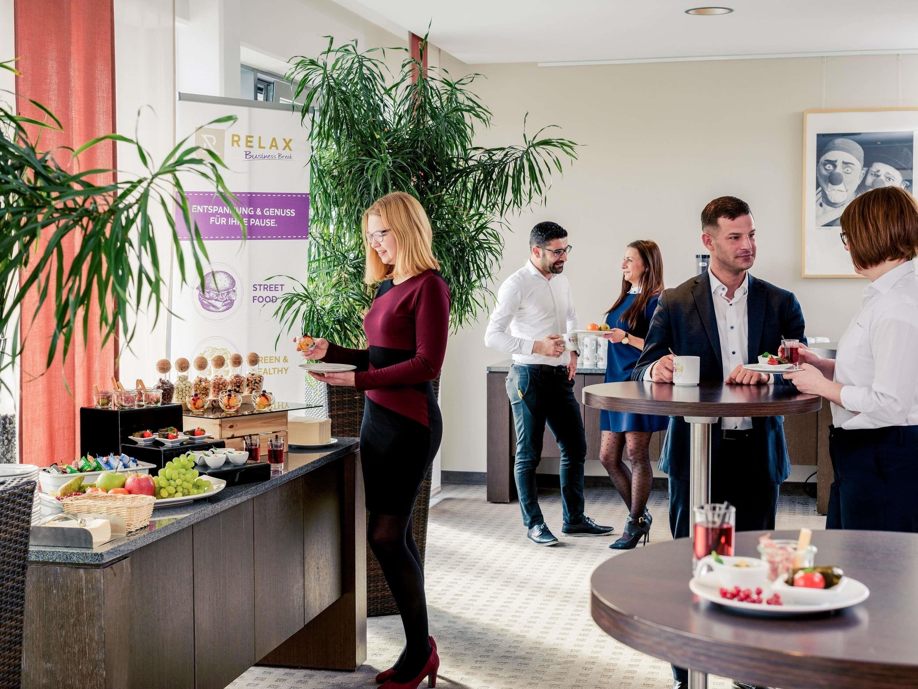 Un espacio interior moderno con una zona de bar, un bufé con diversas opciones de comida, y un grupo de personas conversando.