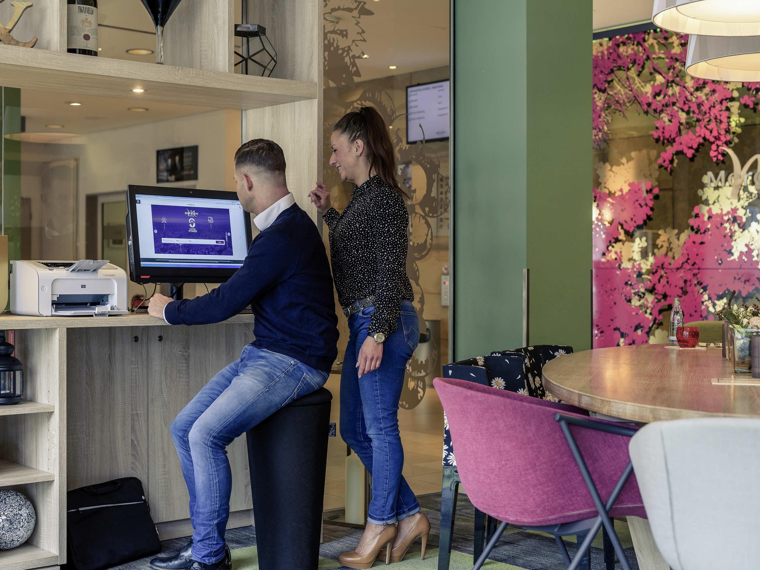 A man and a woman at a reception desk with a computer monitor, a printer, and a glass partition with floral decorations.