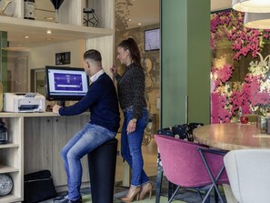 A man and a woman at a reception desk with a computer monitor, a printer, and a glass partition with floral decorations. - Mercure Hotel Hannover Mitte (Hannover)