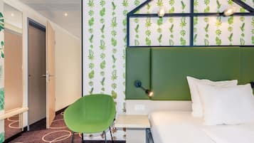 Una habitación de hotel con cabecera verde, una cama con ropa blanca, una silla verde y una pared con estampado de hojas.