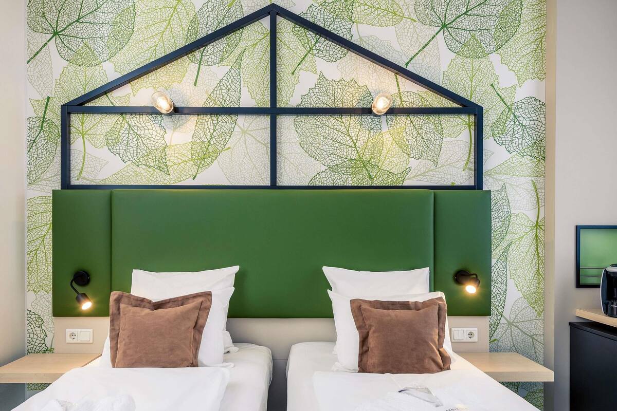 Una habitación de hotel con dos camas, cabeceros verdes, ropa de cama blanca y papel tapiz con estampado de hojas.