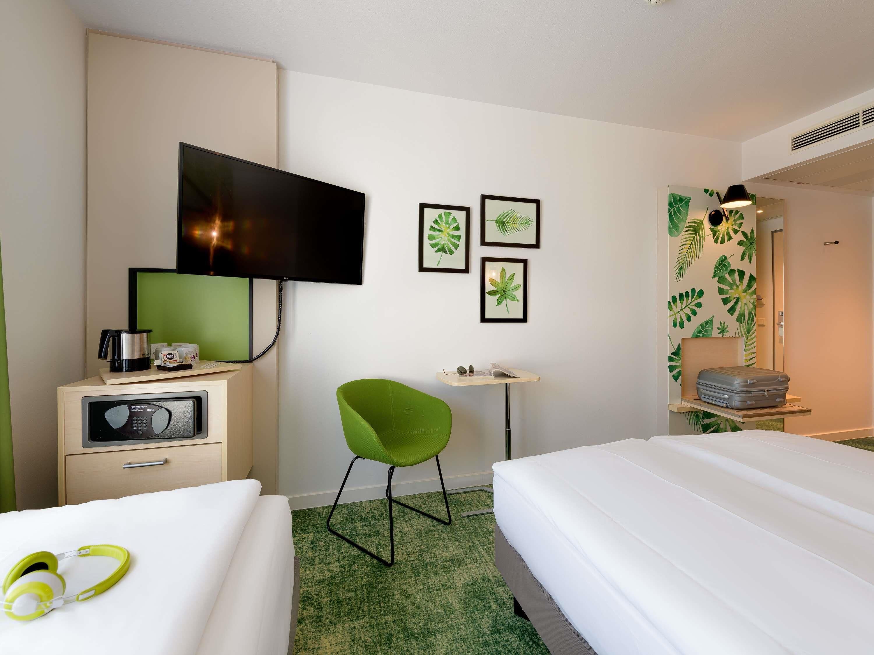 A hotel room with two beds, a desk, a chair, a TV, and a wall with leaf-patterned wallpaper.