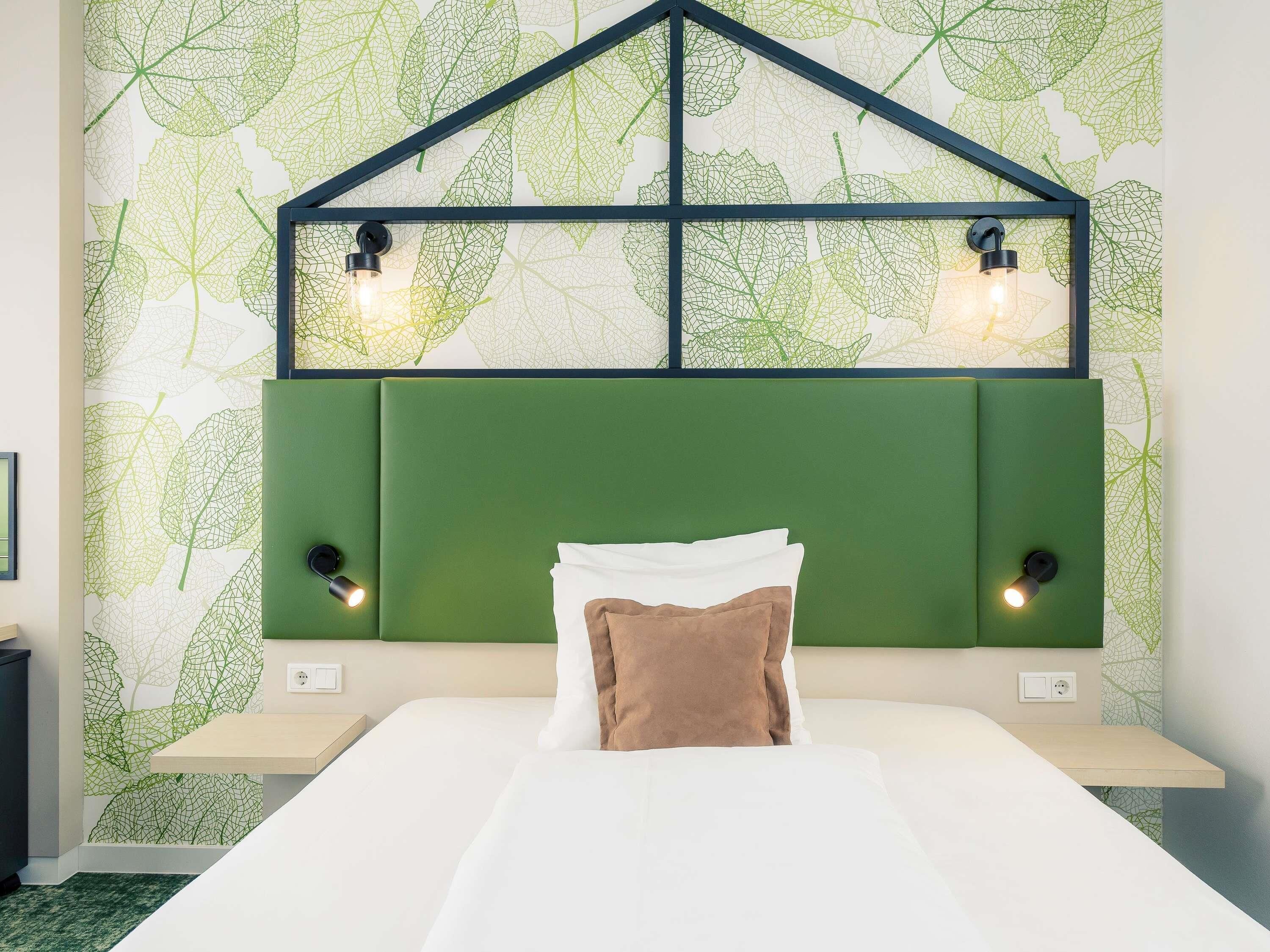 Un dormitorio con cabecera verde, dos lámparas de noche, una cama con ropa de cama blanca y marrón, y una pared con estampado de hojas.