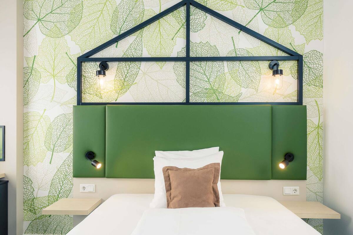 A bedroom with a green headboard, two bedside lamps, a bed with white and brown bedding, and a leaf-patterned wall.