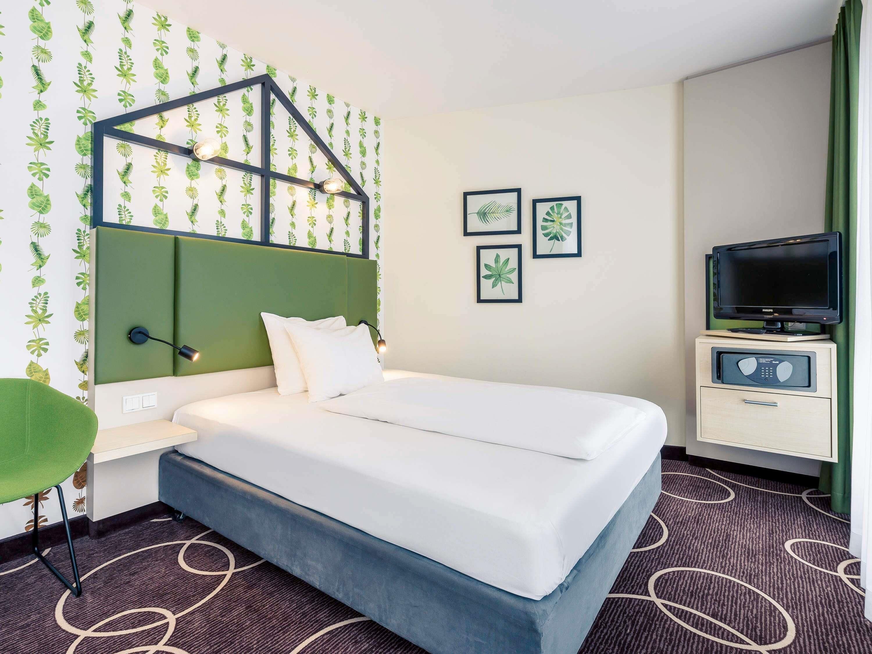 Une chambre d’hôtel avec un lit, une télévision, une table de chevet et un mur recouvert d’un papier peint à motifs de feuilles.