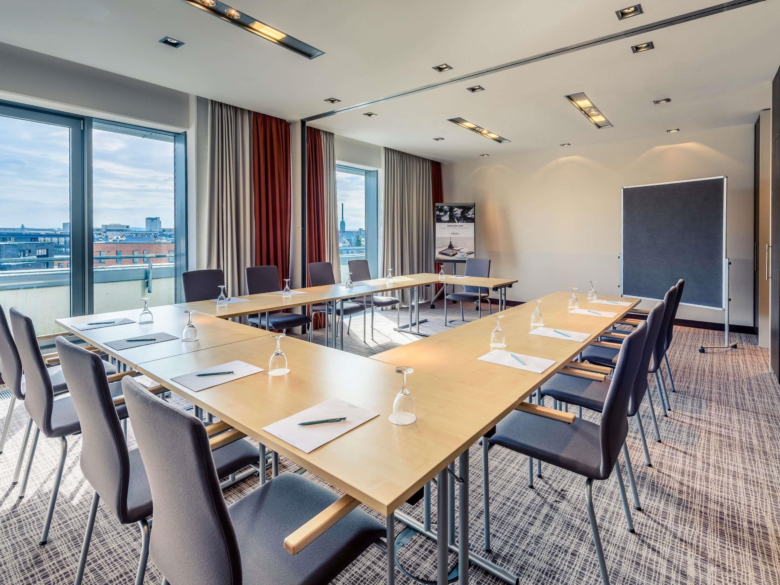 A conference room with a long table, chairs, a projector screen, and large windows with a city view.