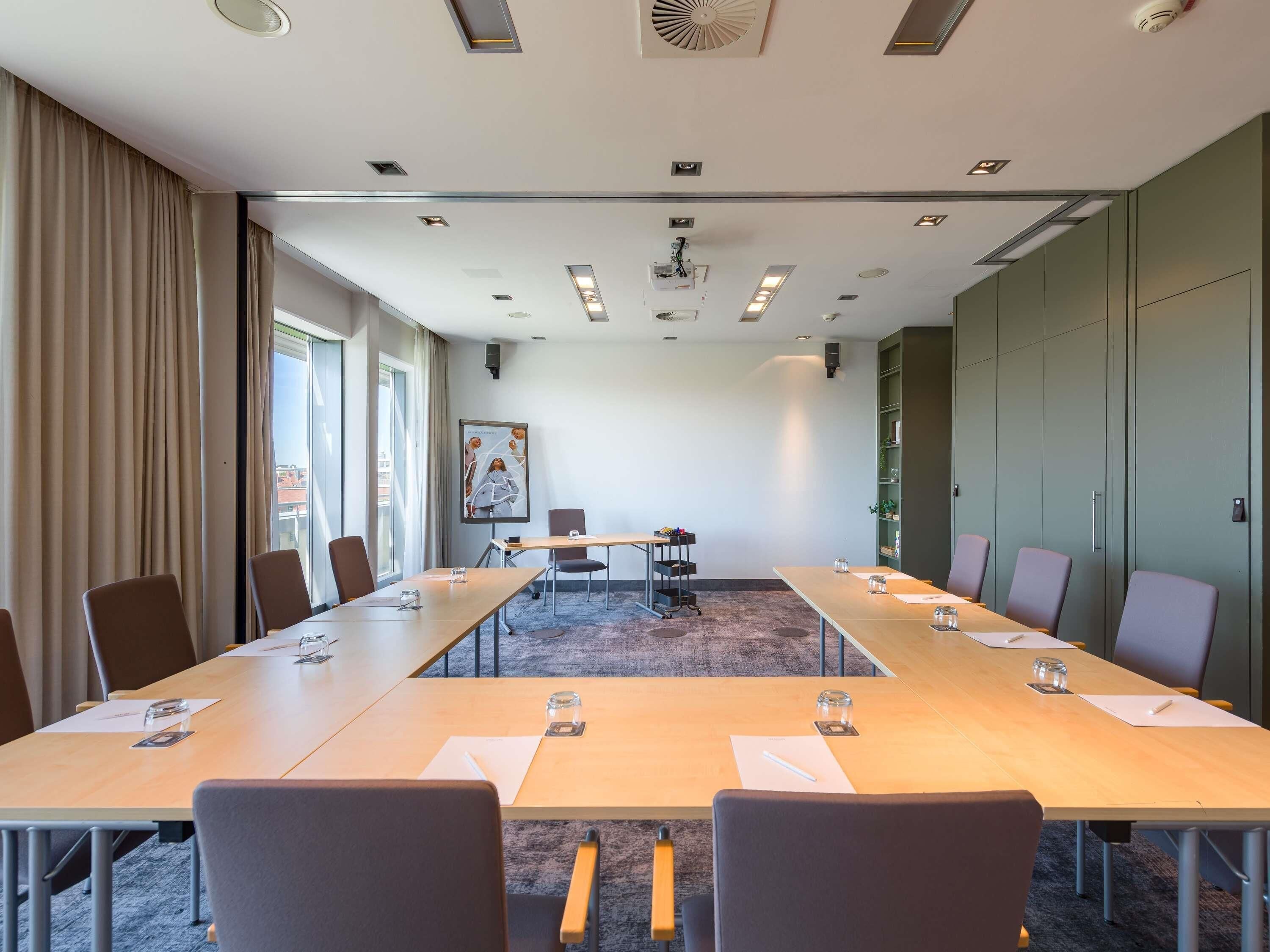 Ein Konferenzraum mit langem Tisch, Stühlen, einer Projektionsleinwand und einem großen Fenster mit Vorhängen.