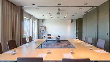 Ein Konferenzraum mit langem Tisch, Stühlen, einer Projektionsleinwand und einem großen Fenster mit Vorhängen.
