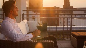 A person sitting on a balcony with a glass of wine, overlooking a cityscape with a prominent church spire. - Mercure Hotel Hannover Mitte (Hannover)