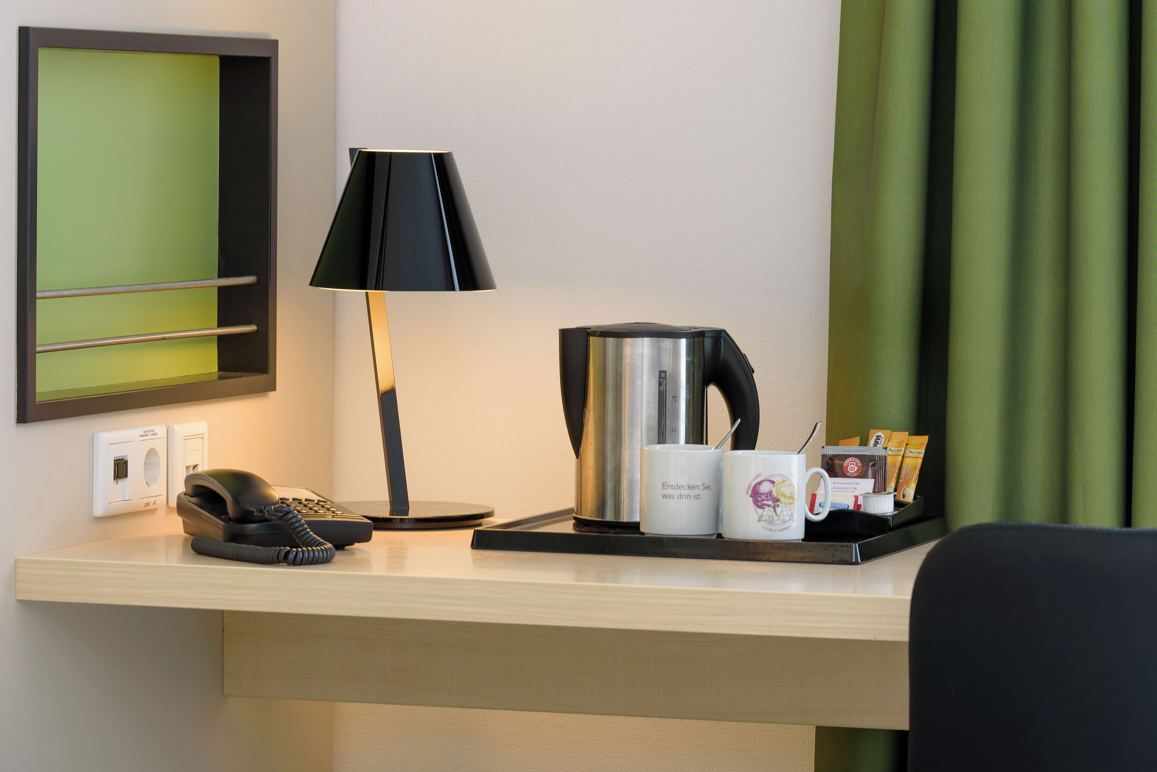 Een hotel bureau met een telefoon, lamp, koffiezetapparaat en een dienblad met een mok en koffiecups.