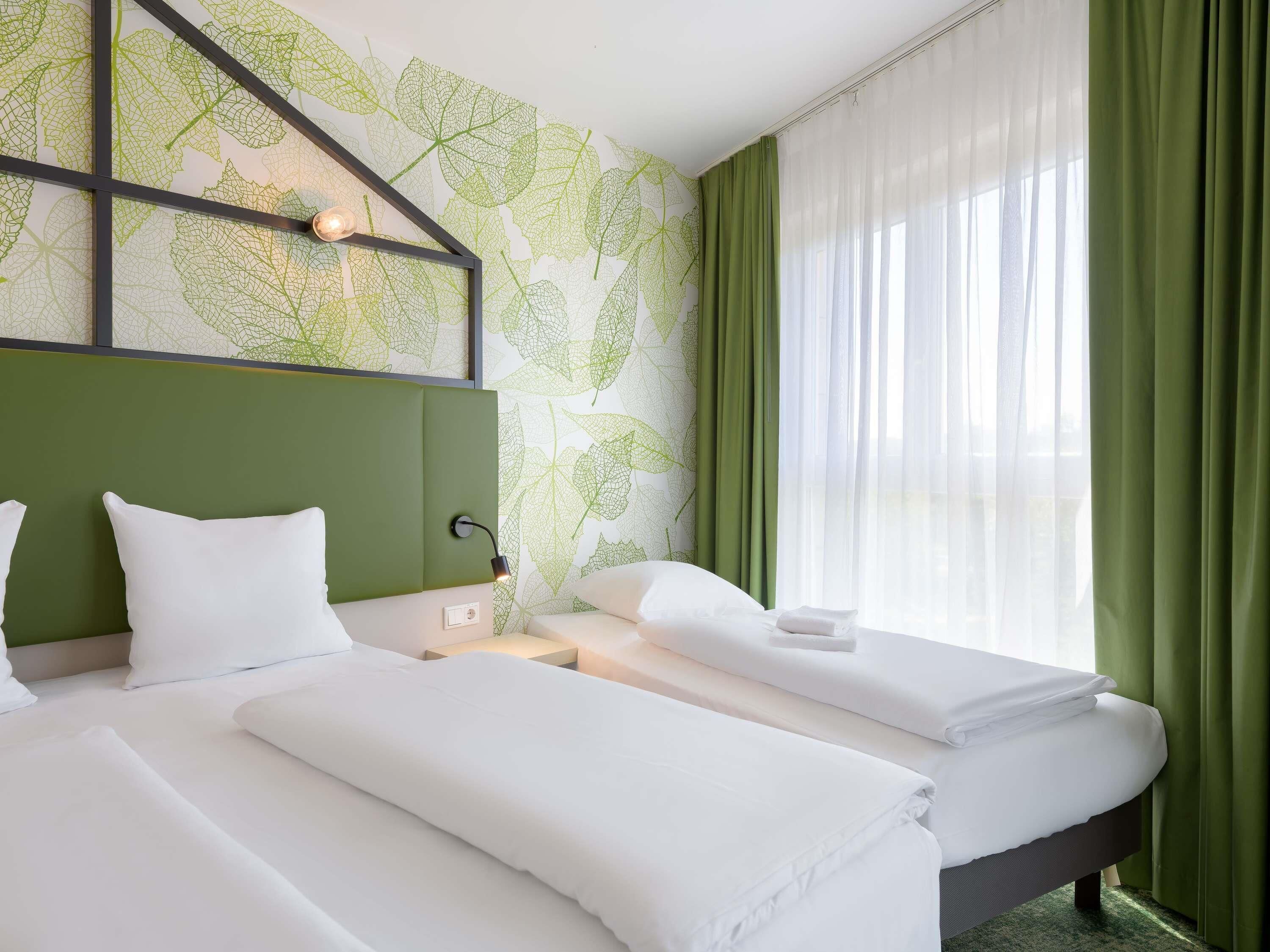 A hotel room with two beds, a green headboard, a wall with leaf patterns, and sheer curtains.