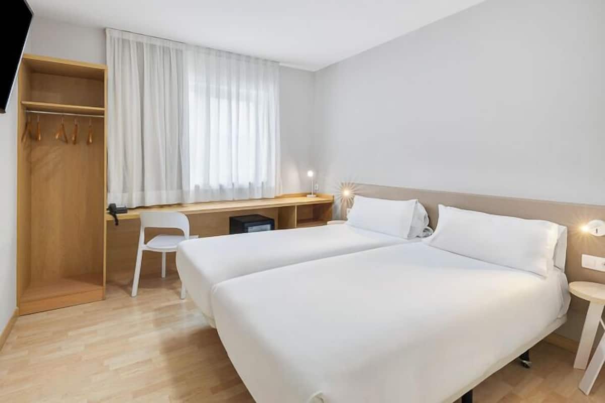A hotel room with two beds, a desk, a chair, a wardrobe, and a window with curtains.