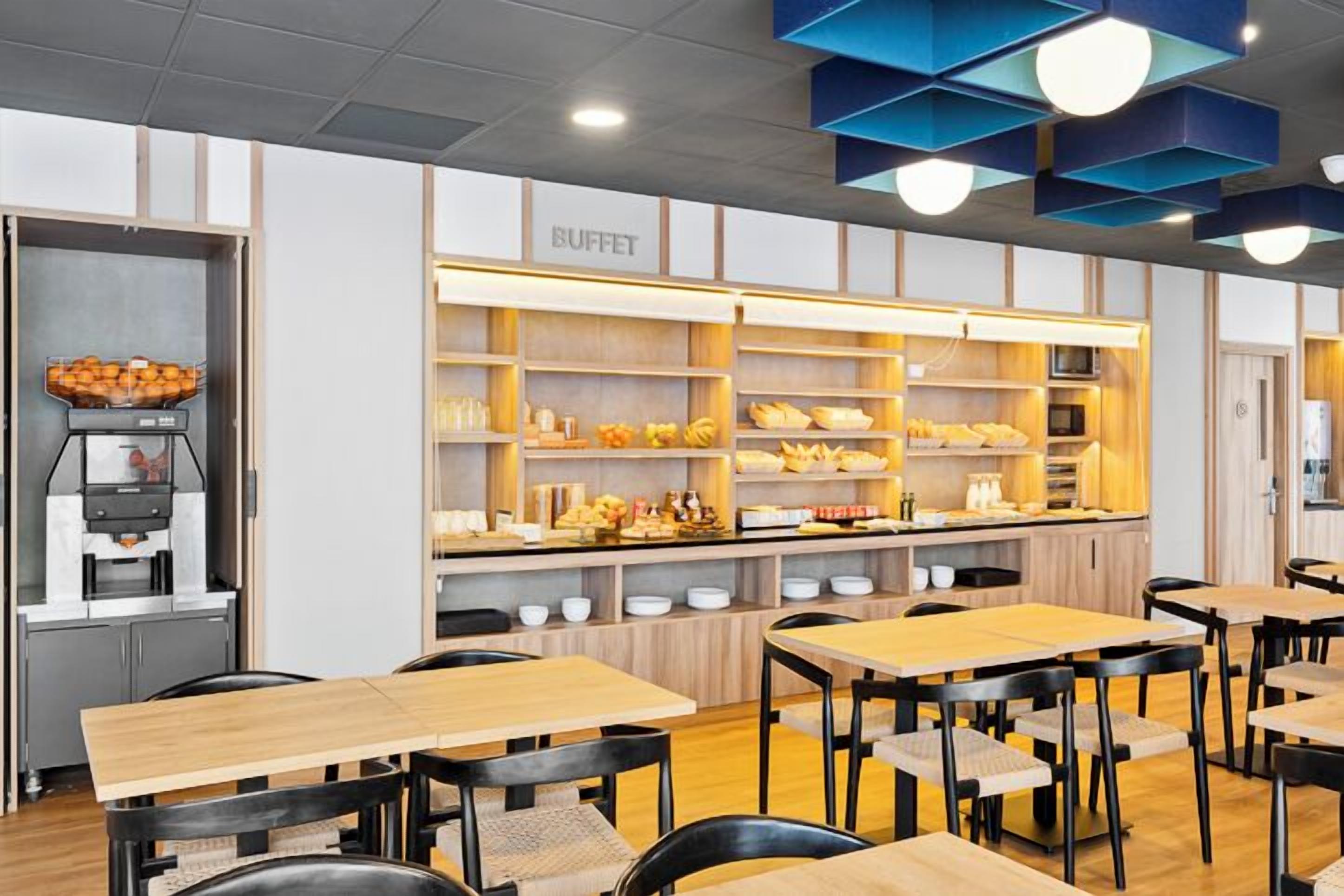 Zona de bufé con mesas y sillas de madera, una máquina de café y diferentes alimentos exhibidos en estantes.