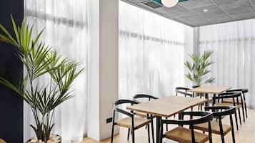 A conference room with a wooden table and chairs, large windows with sheer curtains, and potted plants.
