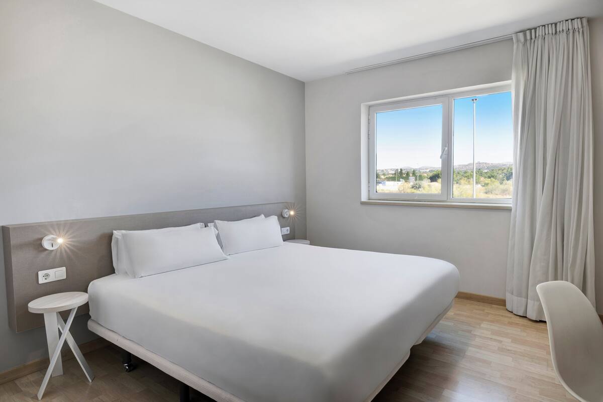 A modern hotel room with a large bed, a small bedside table, a chair, a window with curtains, and a view of the landscape.