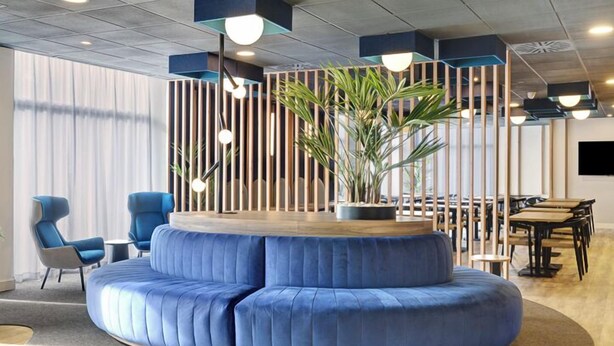A modern lounge area with a circular blue sofa, blue chairs, a wooden table, and a potted plant. - B&B Hotel Las Rozas  (Las Rozas de Madrid)