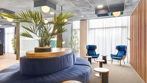 A modern lounge area with a curved blue sofa, round wooden tables, and blue chairs. - B&B Hotel Las Rozas  (Las Rozas de Madrid)