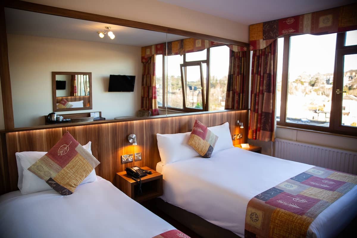 A hotel room with two beds, a bedside table with a phone, a mirror, and a window with curtains.