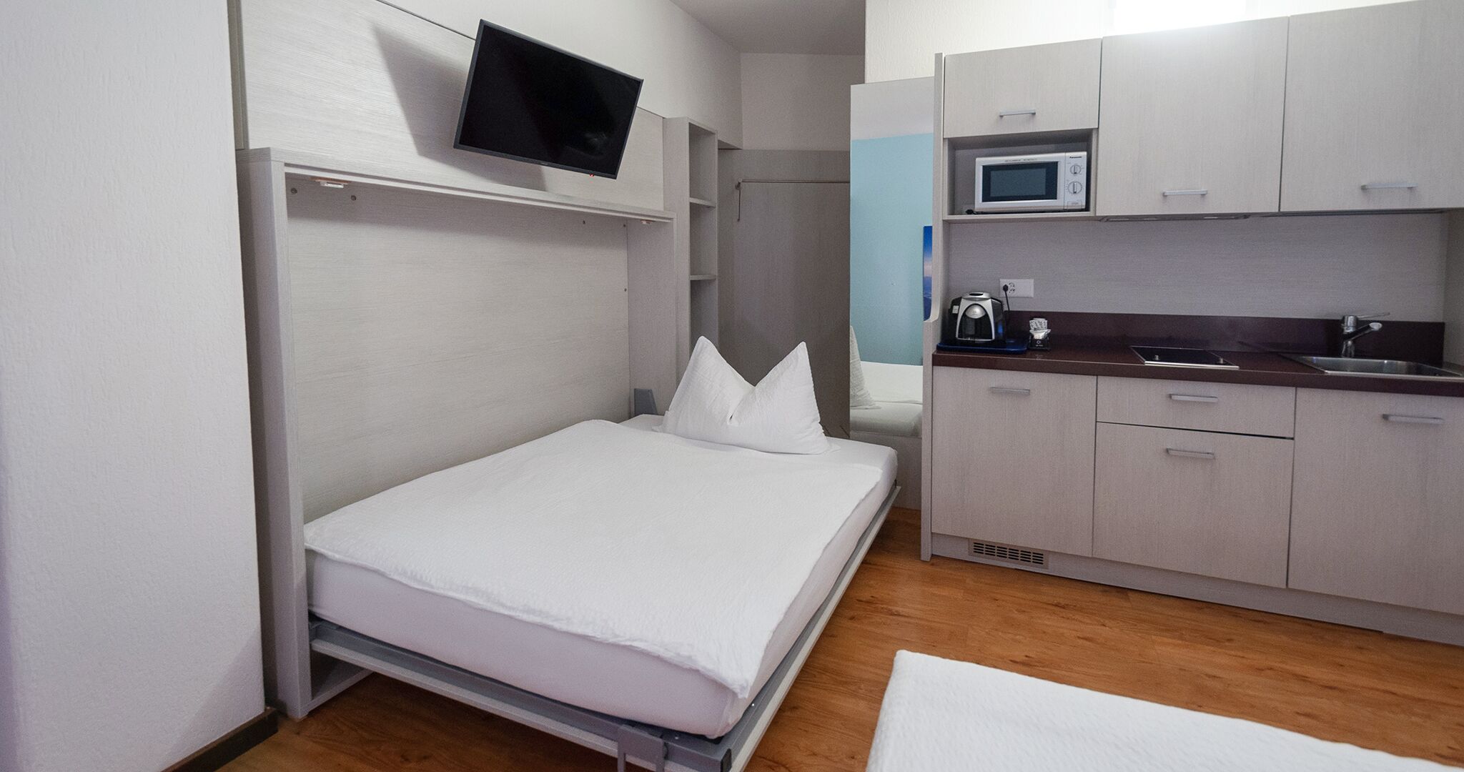Una habitación compacta con una cama, un televisor montado en la pared, un microondas y una pequeña cocina.