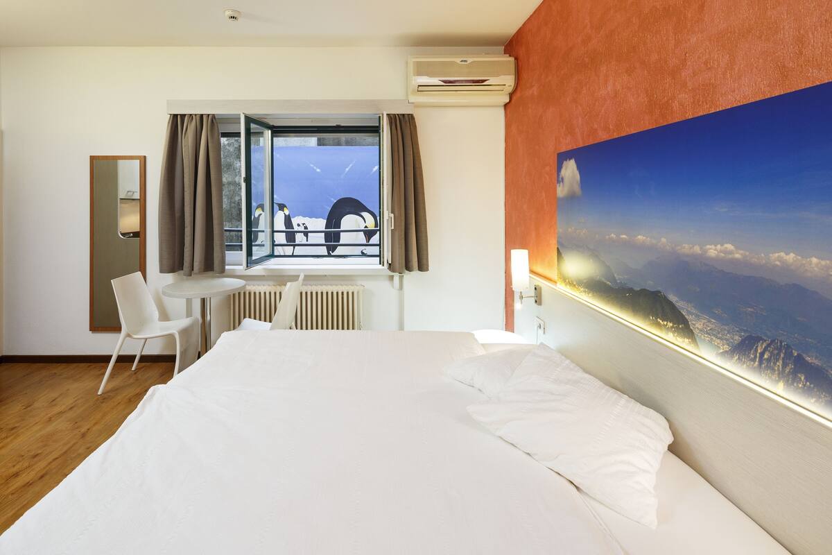 A hotel room with a bed, a desk, a chair, a window with curtains, an air conditioning unit, and a large painting of a mountain landscape.