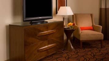 Una camera d'albergo con un armadio in legno, una televisione, una poltrona con cuscino rosso, un tavolino con una lampada e fiori, e un tappeto decorato.