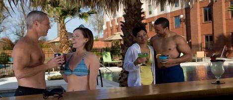 A group of people socializing at an outdoor bar area with a pool and palm trees in the background.