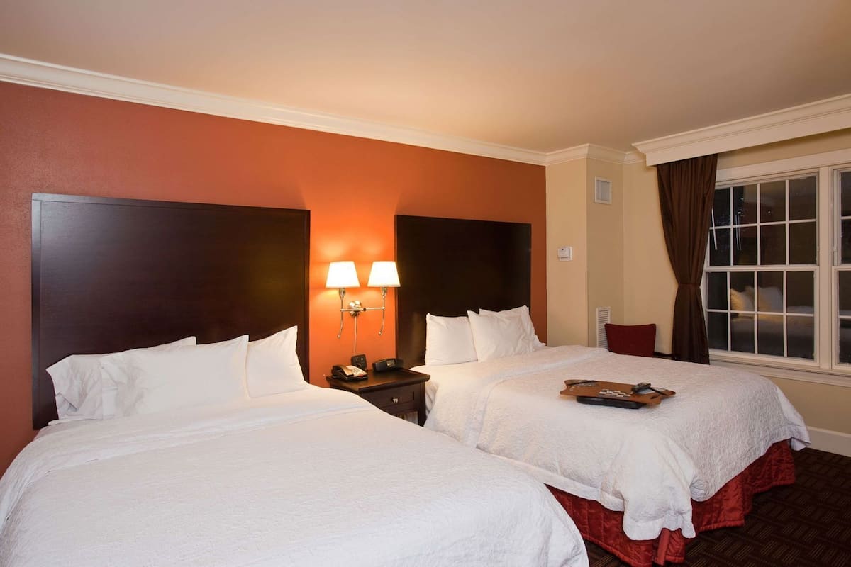 A hotel room with two beds, a nightstand with a lamp, a window with curtains, and a view of the night sky.