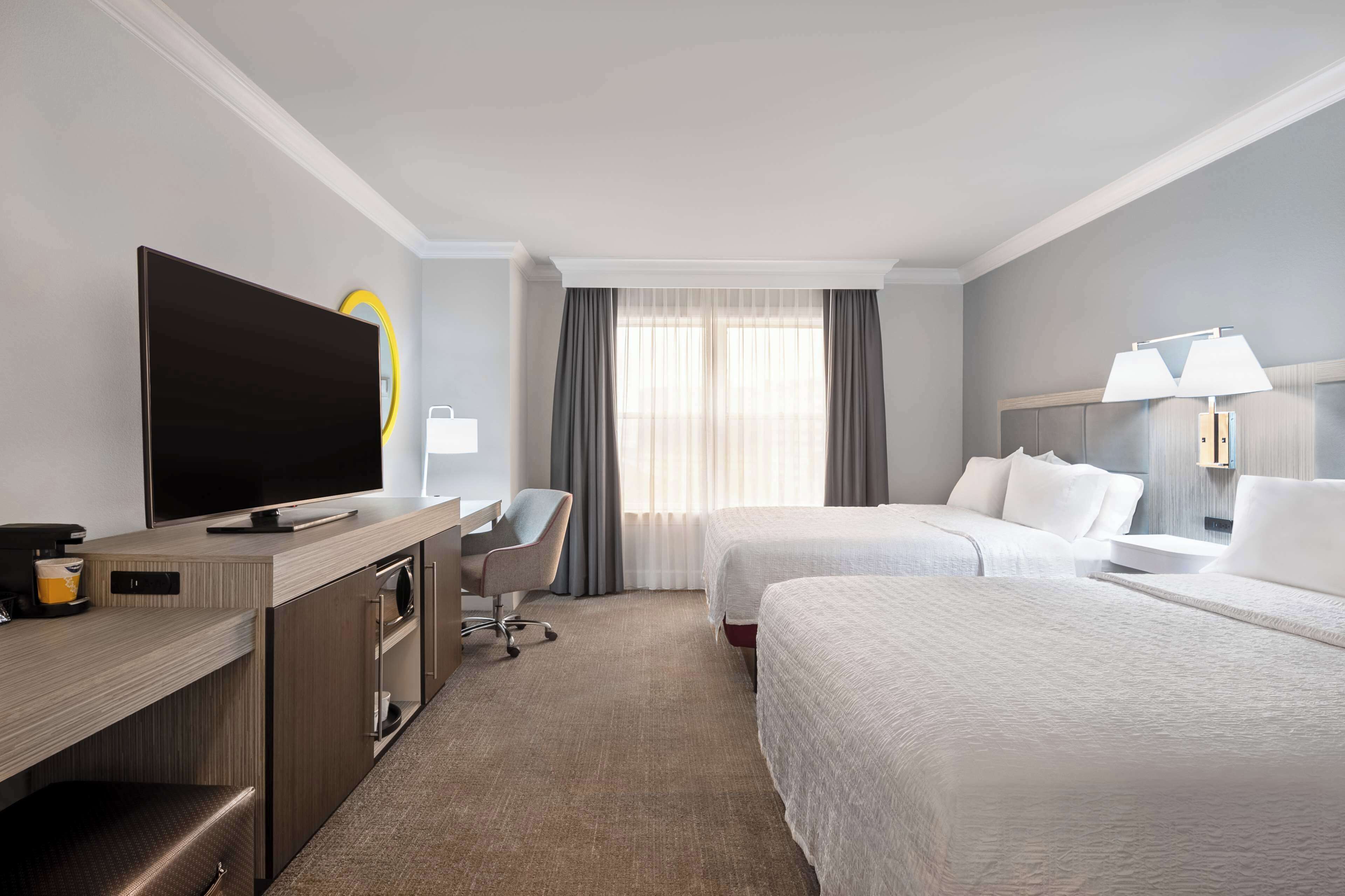 Ein Hotelzimmer mit Flachbildfernseher, Schreibtisch mit Stuhl, zwei Betten, einem Fenster mit Vorhängen und einer Nachttischlampe.
