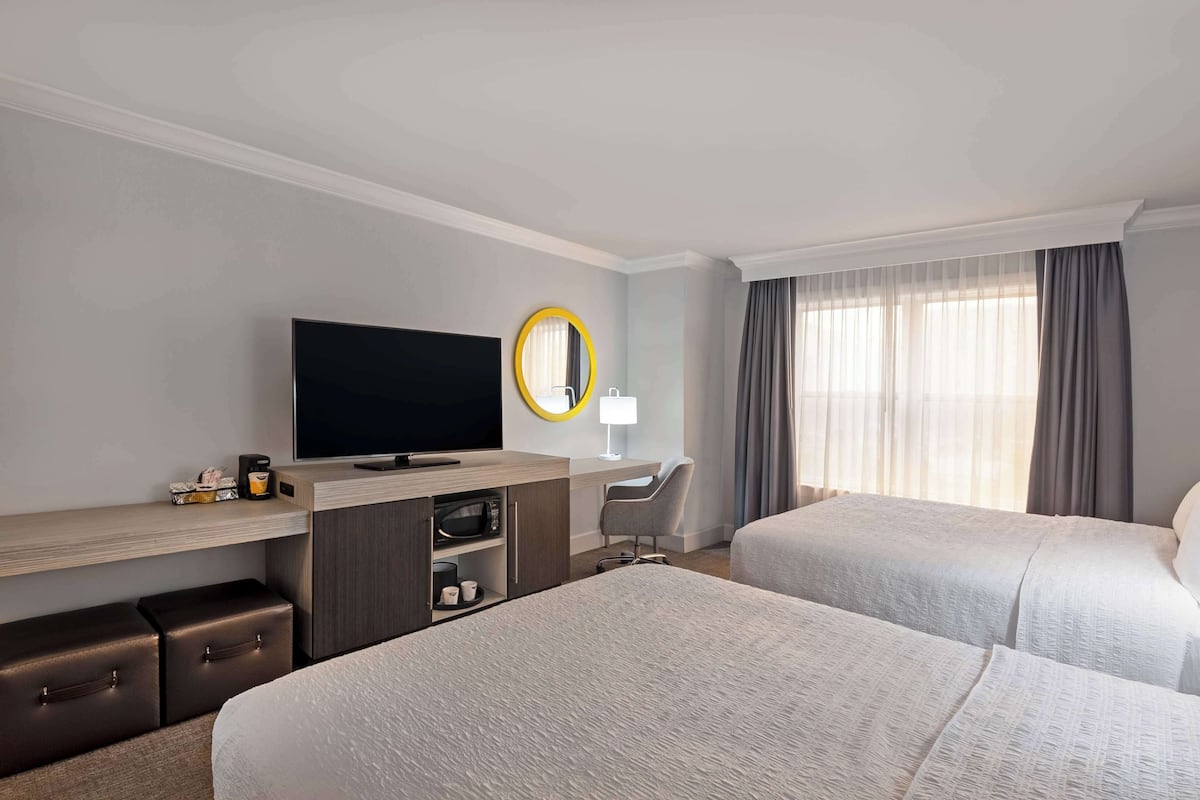 A hotel room with two beds, a TV, a desk with a lamp, a mirror, and a window with curtains.