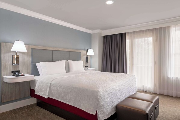A hotel room with a large bed, two bedside tables with lamps, a red accent wall, and a view of a building through sheer curtains.