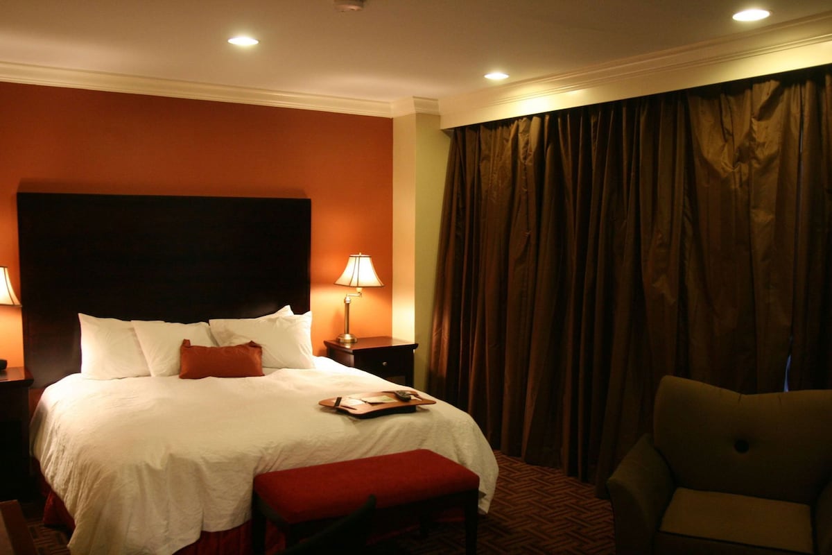 A hotel room with a large bed, two bedside lamps, a red bench, a chair, and a curtain.