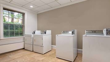 A laundry room with three stacked washers and dryers, a window with a view of greenery, and beige walls.