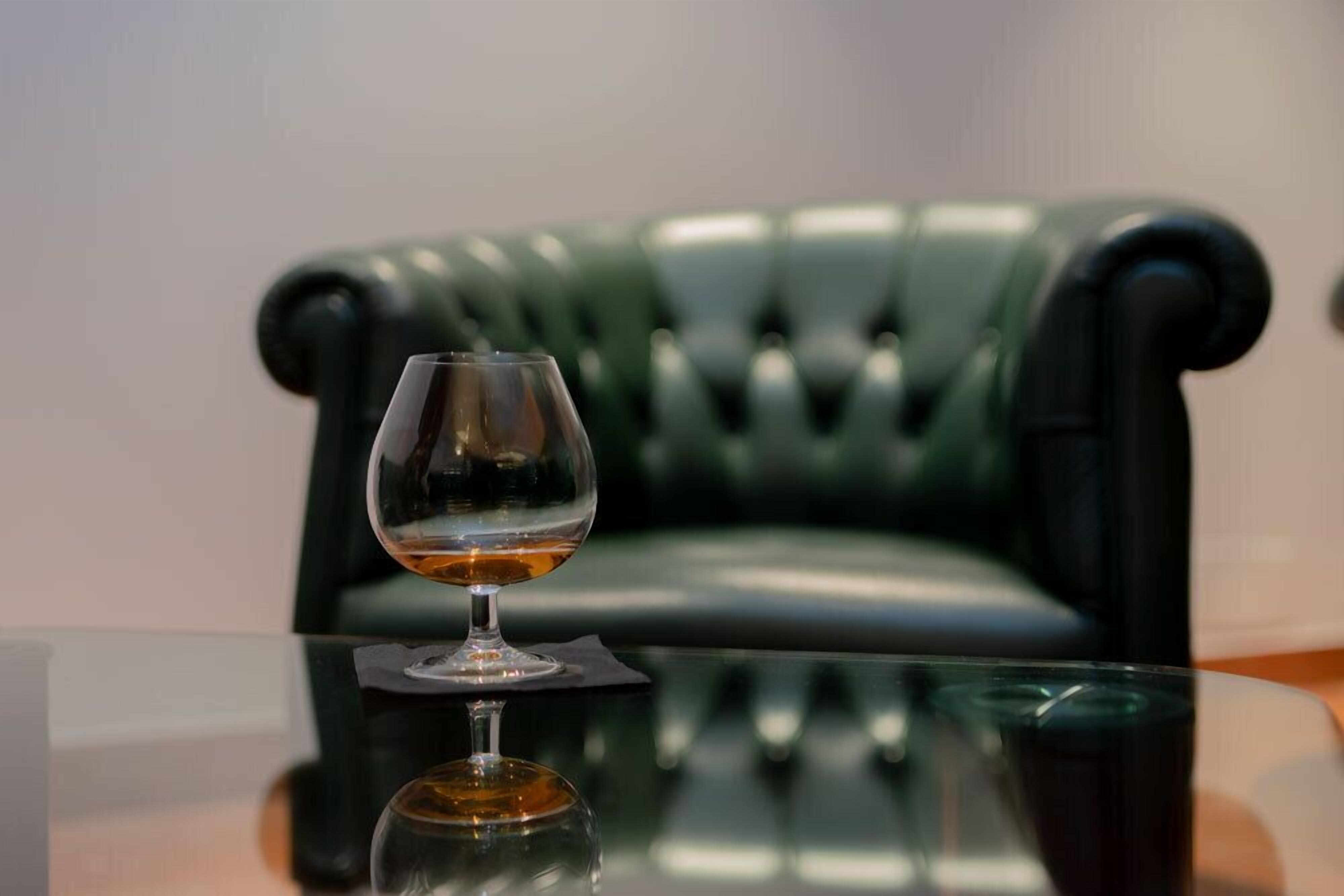 A glass of amber liquid on a reflective surface, with a green upholstered chair in the background.