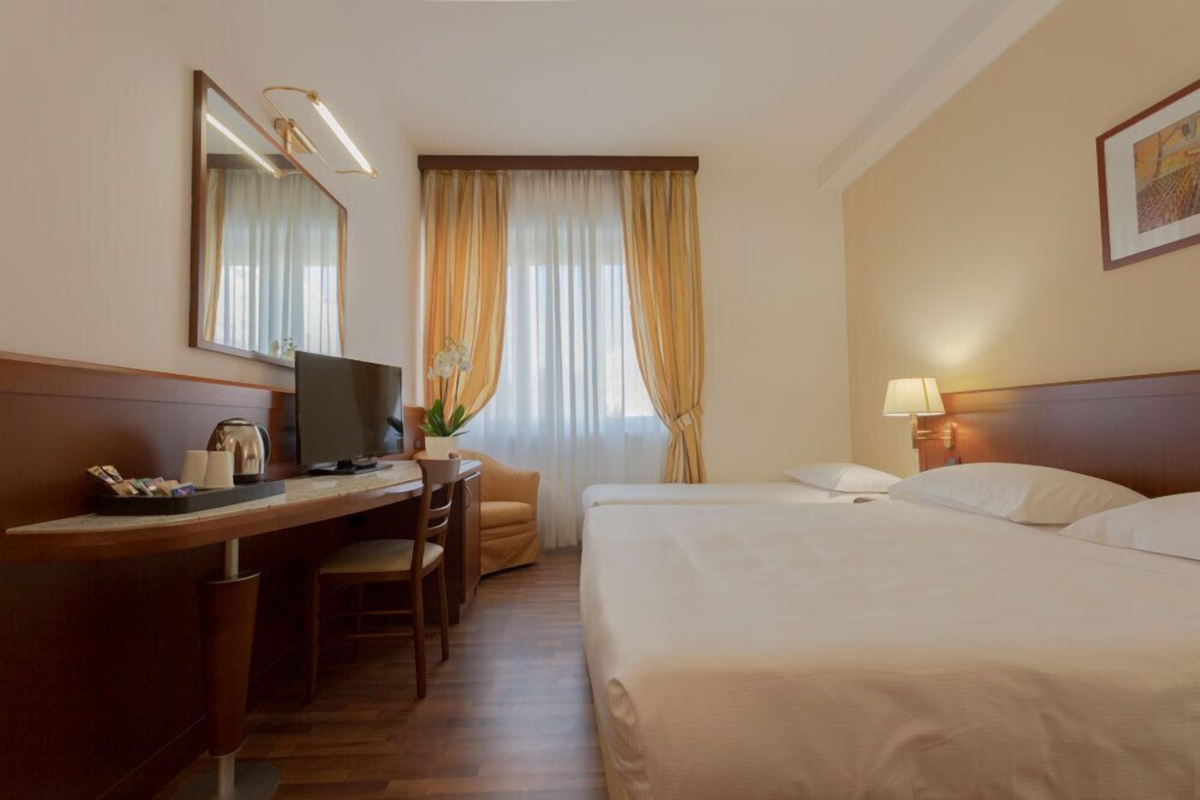 A hotel room with a bed, a desk with a computer, a chair, a lamp, and a window with curtains.