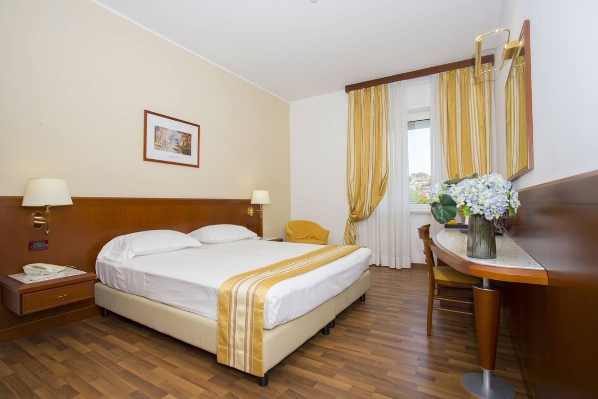 A hotel room with a bed, bedside tables, a desk, a chair, a mirror, and a window with curtains.