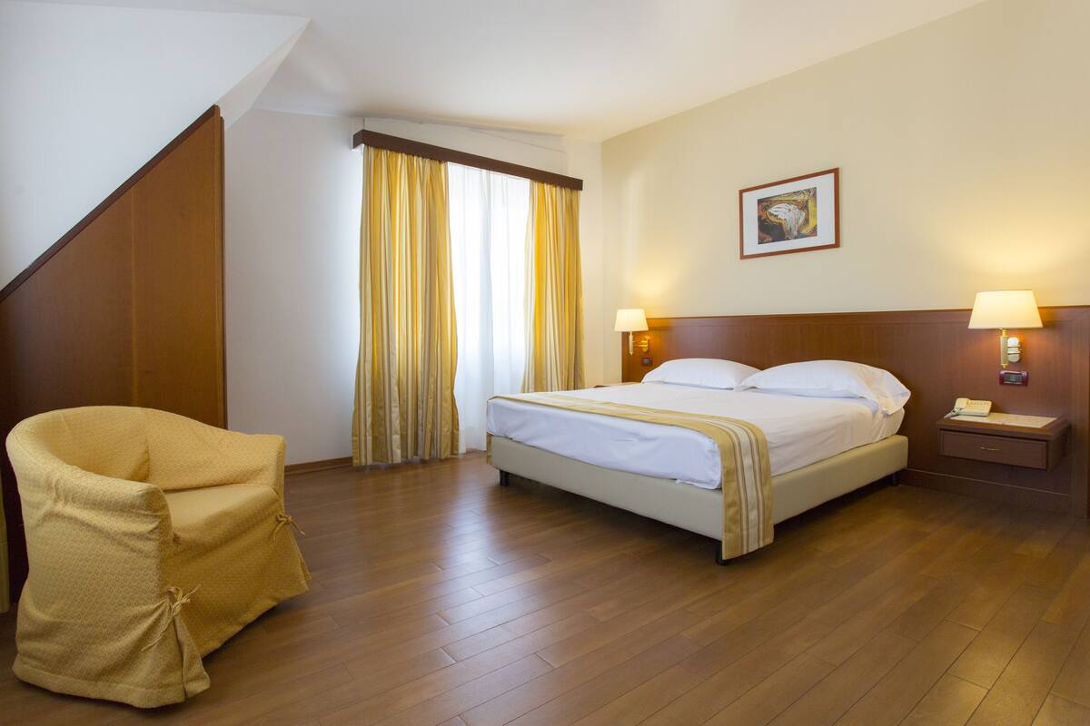 A hotel room with a bed, a chair, a nightstand, and a painting on the wall.