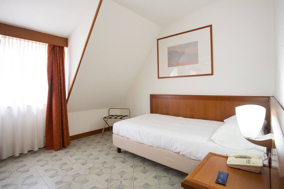 A hotel room with a bed, a bedside table with a lamp, a telephone, and a small table with a packet.